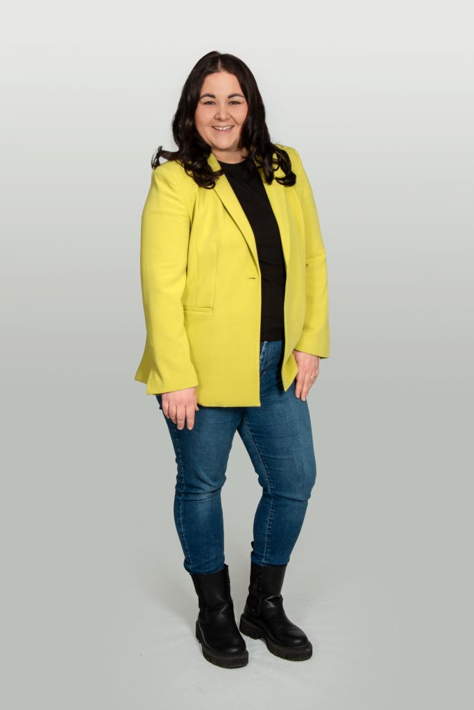 Lisa Bavosa steht vor hellem Hintergrund mit gelbem Blazer und Jeans
